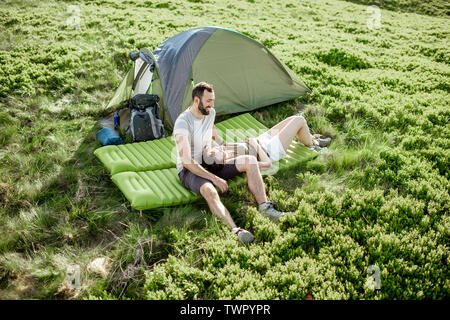 Young lovely couple sitting together on the mattress at the campsite, enjoying summer time while traveling in the mountains Stock Photo