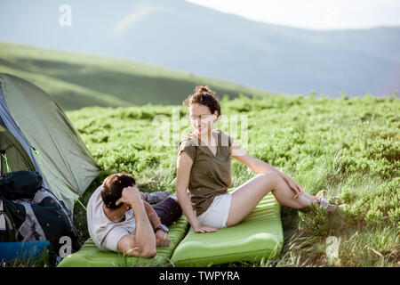 Young lovely couple sitting together on the mattress at the campsite, enjoying summer time while traveling in the mountains Stock Photo