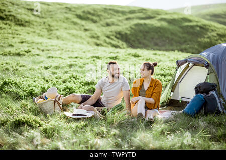 Young and cheerful couple having a picnic at the campsite while traveling high in the mountains during the sunset Stock Photo