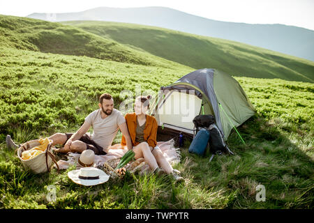 Young and cheerful couple having a picnic at the campsite while traveling high in the mountains during the sunset Stock Photo