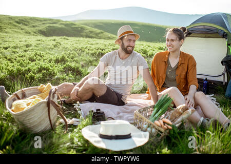 Young and cheerful couple having a picnic at the campsite while traveling high in the mountains during the sunset Stock Photo