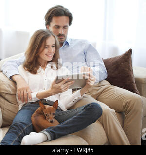 Happy couple using digital tablet at home sitting together with dog on sofa Stock Photo