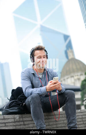 Urban man on smart phone wearing headphones listening to music or having video chat conversation sitting outside using app on 4g smartphones. Casual young urban professional male in Hong Kong Central. Stock Photo