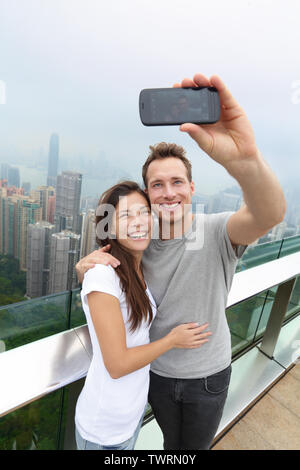Hong Kong Victoria Peak tourists couple taking selfie photo picture with smartphone enjoying view over Hong Kong and Victoria Harbour. Young happy multiethnic couple traveling in Asia. Stock Photo