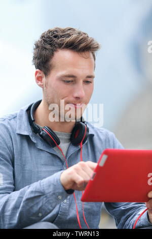 Urban young professional man using tablet computer sitting outside using app on 4g wireless device wearing headphones. Casual young urban professional male in his late 20s. Stock Photo