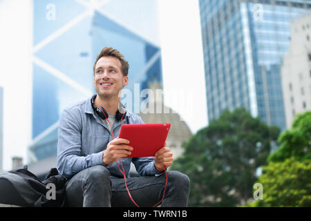 Urban man using tablet computer sitting in Hong Kong outside using app on 4g wireless device wearing headphones. Casual young urban professional male in his late 20s. Hong Kong Central. Stock Photo