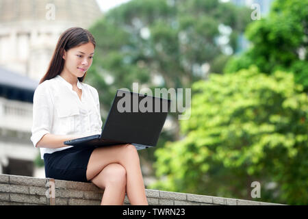 Business woman with computer laptop working outside looking at screen in business district, Central, Hong Kong. Young female professional businesswoman sitting outdoors. Asian Chinese Caucasian woman. Stock Photo