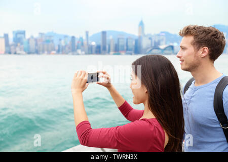 Tourist couple taking photo pictures enjoying view and sightseeing on Tsim Sha Tsui Promenade and Avenue of Stars in Victoria harbour, Kowloon, Hong Kong. Tourism travel concept. Stock Photo