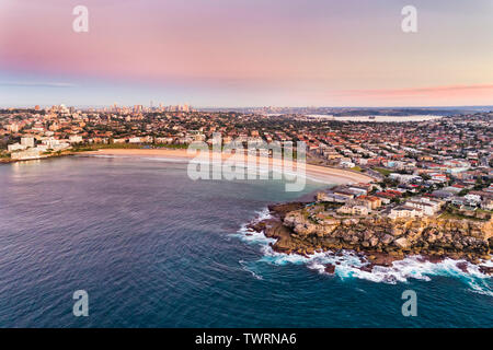North Bondi sandstone cliff of headland guarding famours Bondi Beach of Sydney from open waves of Pacific ocean in aerial sunrise view towards distant