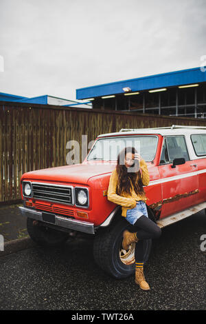 Young happy woman in orange sweater and jeans short near old retro red american car. Hitchhiking hippie travel concept. Autumn look. Stock Photo