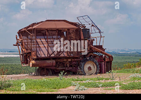 a broken down rusty old cotton picking machine sits above modern fields and greenhouses in israel with a palestinian city in the background Stock Photo