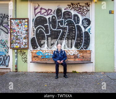 Senior, elderly man sitting on graffiti-covered wooden Bench in front of blind covered in graffiti tags in Friedrichshain-Berlin Stock Photo