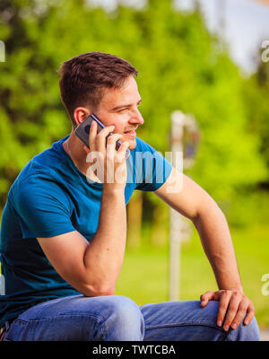 Handsome young man is sitting outdoors and talking with someone on the phone Stock Photo