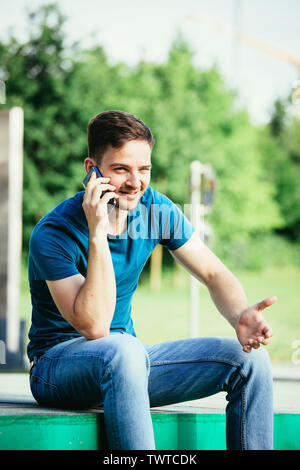 Handsome young man is sitting outdoors and talking with someone on the phone Stock Photo