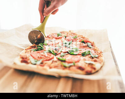high angle view of hand cutting delicious homemade pizza Margherita with tomatoes and basil using a pizza cutter Stock Photo