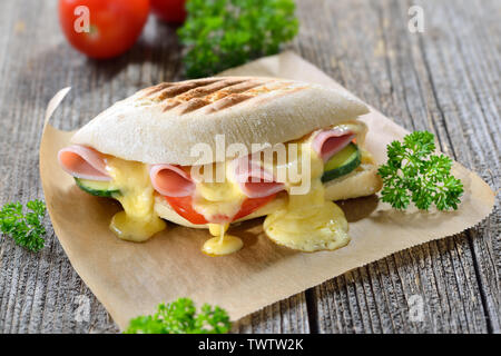 Grilled and pressed panini with ham, melted cheese, cucumber and tomato served on sandwich paper on a wooden table Stock Photo