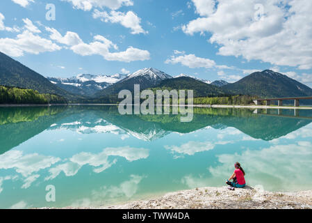 Woman sitting on the shore of the Sylvenstein reservoir and looking at the Karwendel mountains reflected in the turquoise water, Bavaria, Germany Stock Photo