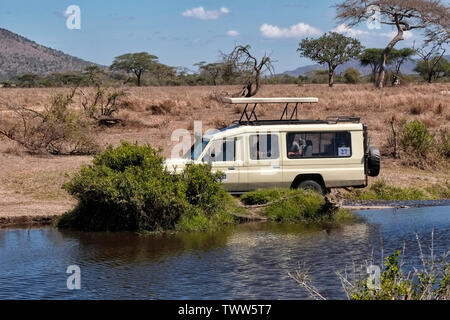 Tourists in a closed 4WD Toyota Landcruiser safari vehicle with a pop-top roof crossing a waterway at the Serengeti National park a UNESCO World Heritage Site in Tanzania Eastern Africa Stock Photo