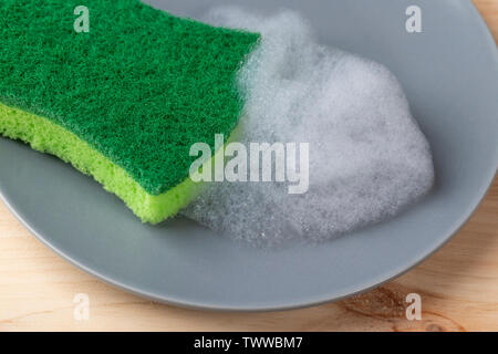A hand with a green spunge washes a gray ceramic plate with detergent foam. On a wooden table. Stock Photo