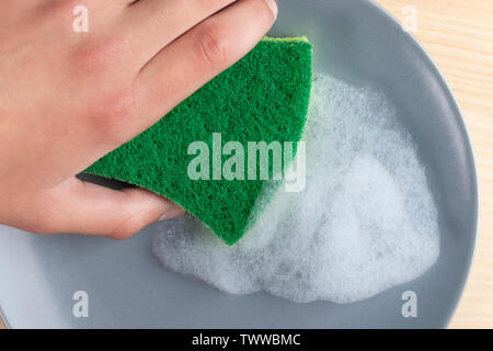 A hand with a green spunge washes a gray ceramic plate with detergent foam. On a wooden table. Stock Photo