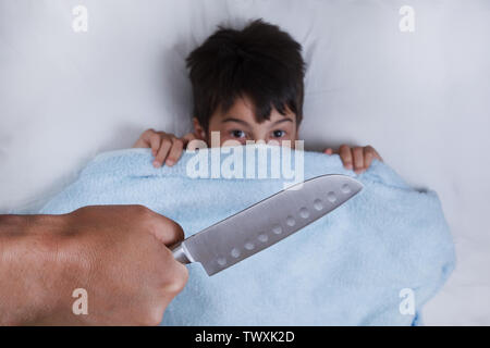 the knife in a hand, against the background of the child hid under a blanket for fear, the concept of child domestic violence the top view Stock Photo