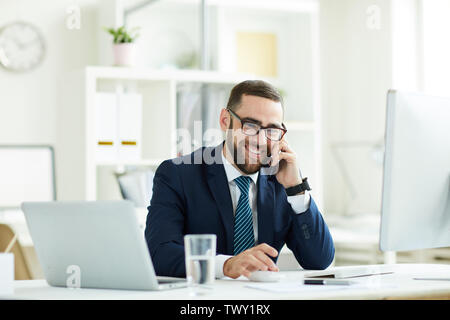 Cheerful excited young bearded manager in eyeglasses sitting at desk with modern computers and talking on mobile phone Stock Photo