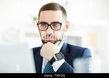 Serious pensive young bearded businessman in glasses concentrated on work looking at computer screen and analyzing online data Stock Photo
