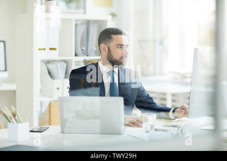 Serious focused young bearded business analyst in suit sitting at desk and making financial forecast Stock Photo