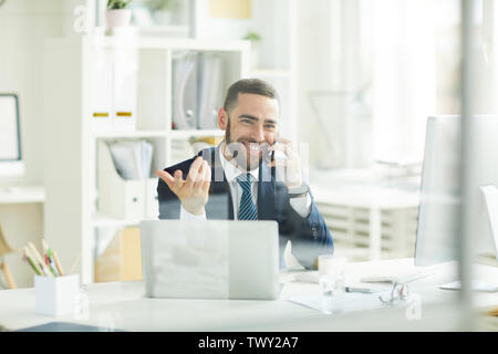 Positive confident young bearded trader in formal suit sitting at table and gesturing hand while consoling customer about financial assets on phone Stock Photo