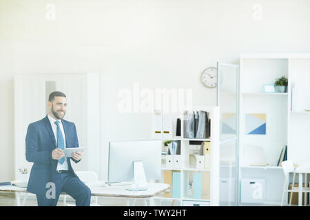 Jolly ambitious handsome young project manager in formal suit sitting on table and using tablet while elaborating strategy Stock Photo