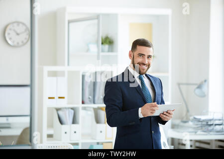 Portrait of cheerful confident handsome young manager in dark suit standing in office and using digital tablet Stock Photo