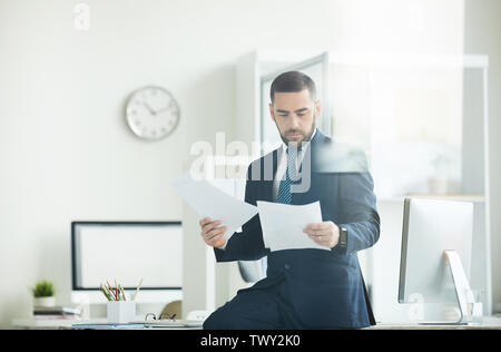 Serious focused young bearded manager in suit sitting on desk and examining papers while doing financial expertise Stock Photo