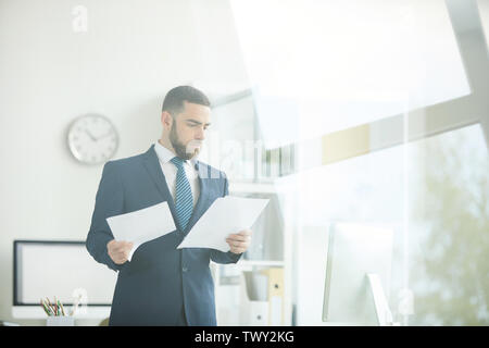Serious busy young bearded businessman in suit standing in modern office and working with business documents Stock Photo