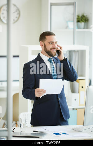 Optimistic handsome young bearded businessman in dark blue suit standing in modern office and having phone conversation while discussing papers with c Stock Photo