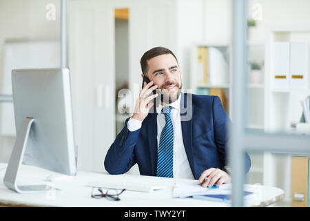 Positive confident handsome young project manager with beard sitting at table and discussing plans with colleague on mobile phone Stock Photo