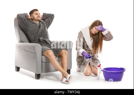 Tired wife doing chores while her lazy husband sitting in armchair against white background Stock Photo
