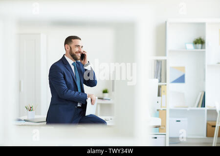 Positive handsome bearded product manager in formal suit sitting on table and using mobile phone while agreeing on product launch Stock Photo