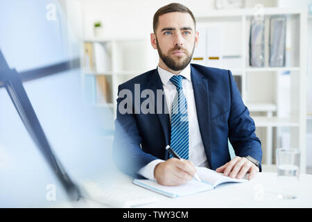 Serious purposeful young bearded businessman in formal suit sitting at table and making notes in diary while planning schedule Stock Photo