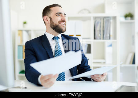Positive excited young bearded financial manager in suit sitting at table in office and analyzing business documents Stock Photo