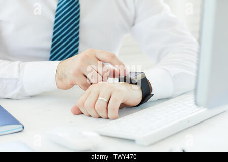 Close-up of unrecognizable businessman in white shirt sitting at desk with computer and reading message on smartwatch Stock Photo
