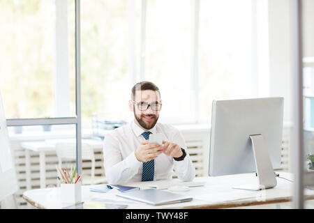 Smiling confident handsome young bearded manager in eyeglasses sitting at table and using smartphone while working in office Stock Photo