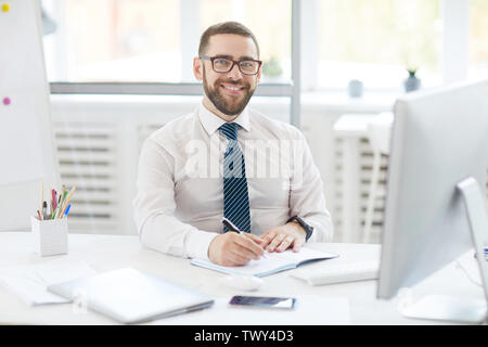 Portrait of positive confident handsome young manager in eyeglasses sitting at table and making notes in organizer Stock Photo