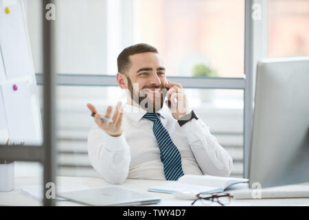Jolly handsome young employee with beard sitting at desk and looking at computer monitor while answering call in office Stock Photo