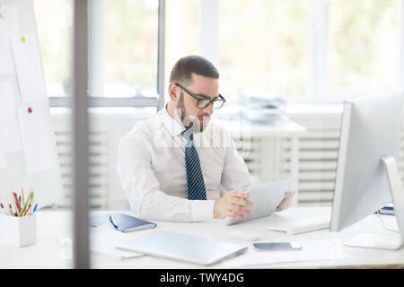 Concentrated young bearded businessman in glasses sitting at desk and analyzing online chart on tablet Stock Photo