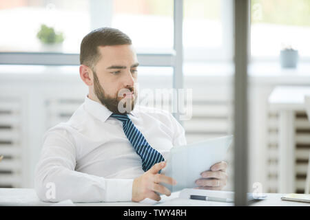 Serious busy young bearded manager sitting in relaxed pose at table in office and using tablet while checking data Stock Photo