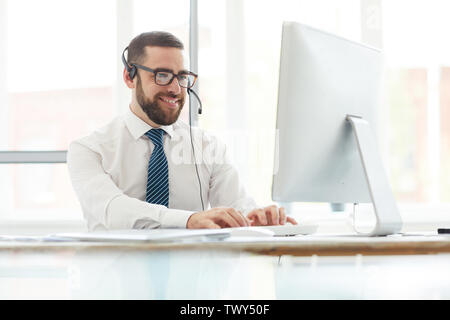Positive handsome young switchboard operator in headset sitting at table and typing on computer keyboard while answering website message of customer Stock Photo