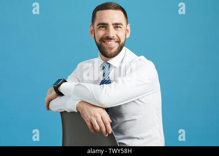 Portrait of cheerful purposeful young bearded manager with smartwatch sitting on chair against blue wall Stock Photo