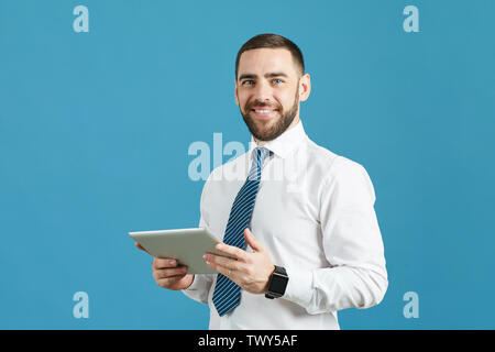 Positive handsome young manager with beard standing against blue wall and using digital tablet while looking at camera Stock Photo