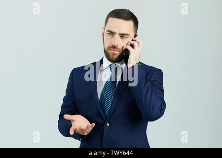 Serious displeased young bearded businessman in formal suit standing against white wall and gesturing hand while talking on mobile phone Stock Photo