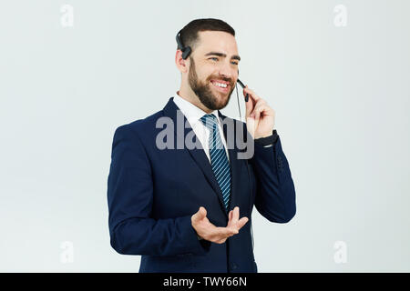 Positive handsome young bearded call center operator in suit standing against white wall and gesturing hand while answering customer on headset Stock Photo